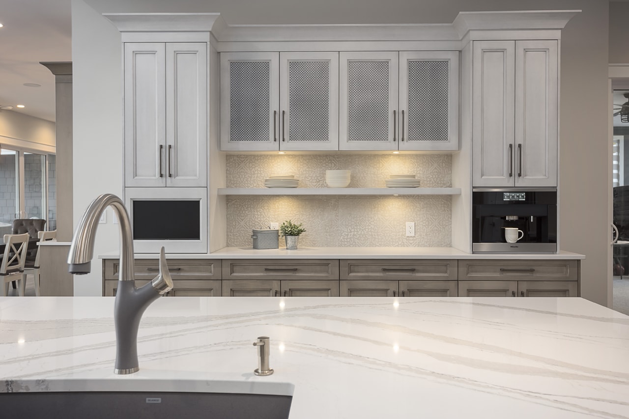 Looking over a white quartz kitchen island onto white brush-stroked glazed cabinets with wire inserts and a single floating shelf with dishes.